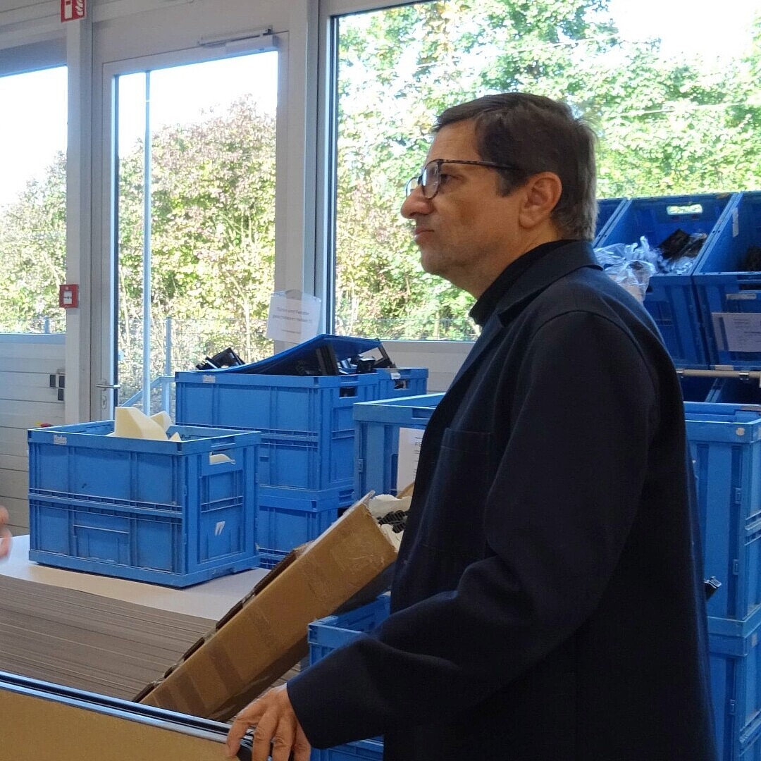 Two men (Konstantin Thomas and Rudi Raschke) stand in a warehouse with shelving and blue plastic bins, one holding a cardboard box and the other gesturing with his hand.