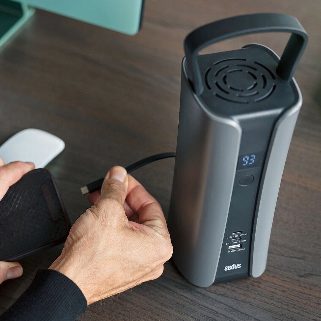 Hands holding a USB-C cable near a portable battery labeled 'se:charge' on a wooden desk with a keyboard and mouse nearby