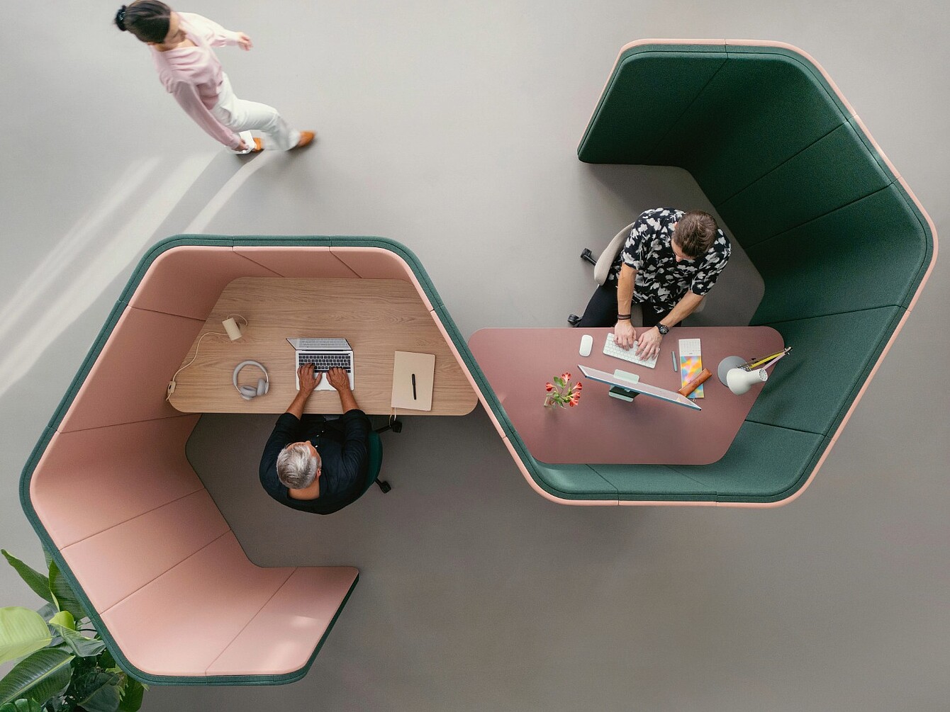 Due persone che lavorano separatamente a scrivanie curve con divisori imbottiti alti in un ufficio open space, una che digita su un laptop e l’altra che scrive su carta, mentre una terza persona cammina nelle vicinanze.