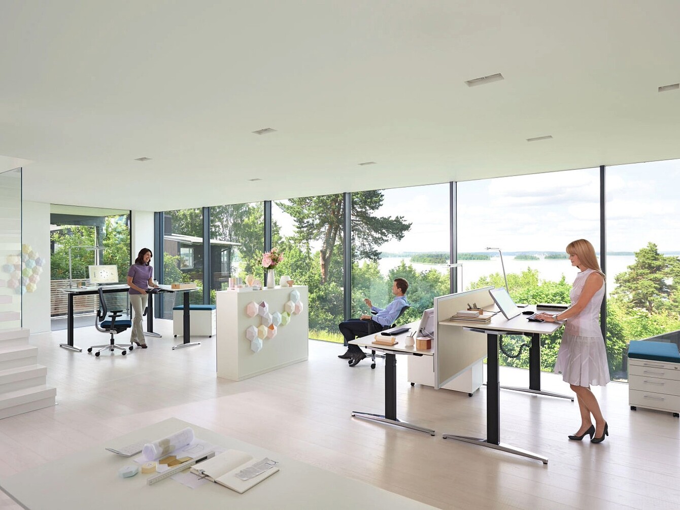 Moderno spazio ufficio con ampie finestre che offrono una vista sul verde esterno, con tre persone che utilizzano scrivanie regolabili in altezza in posizione seduta e in piedi, con mobili bianchi minimalisti e pavimenti chiari.