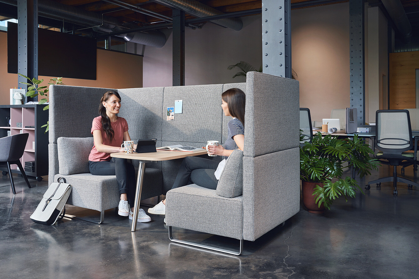 Due donne sedute su una panca grigia dallo schienale alto in un ufficio open space, intente a conversare con delle tazze in mano