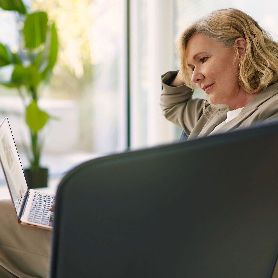 Person with shoulder-length hair sitting in a curved armchair using a laptop with a plant and a cup on a table nearby