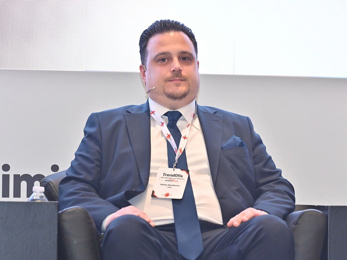 Uomo in giacca e cravatta seduto su una poltrona nera con un badge da conferenza con la scritta “TrendOfis”