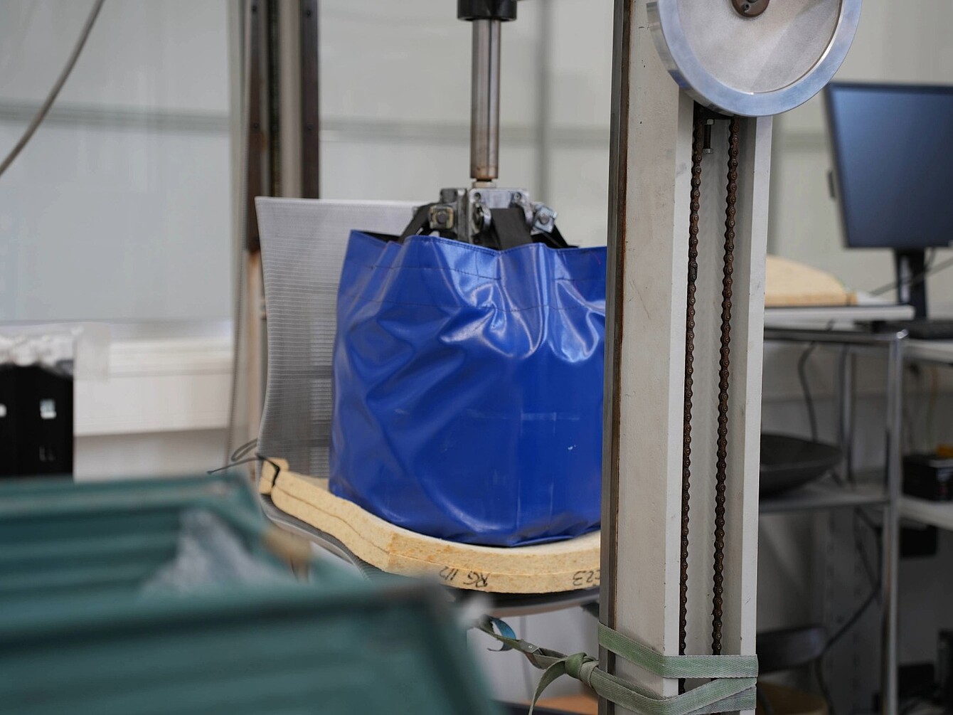 Bolsa azul suspendida en un dispositivo de ensayo mecánico con base de madera y estructura metálica en un entorno de laboratorio
