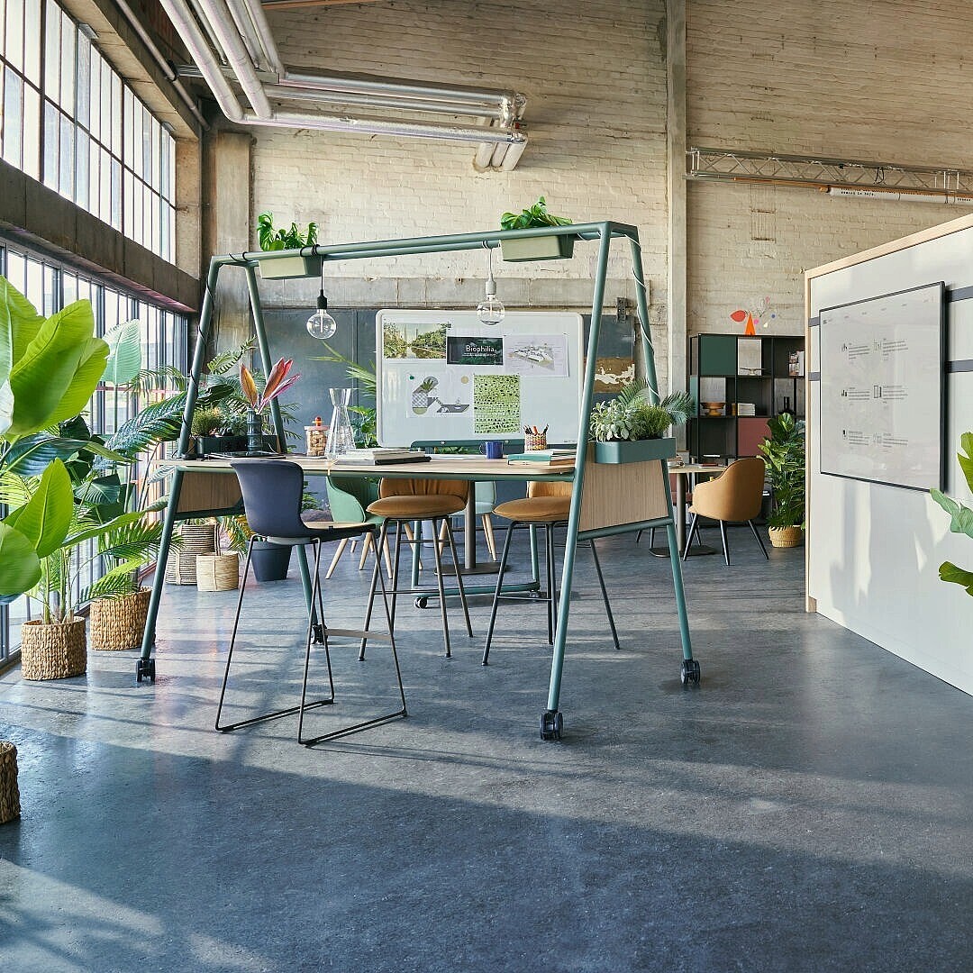 Open-plan office space with high tables and stools, whiteboards, plants, and natural light from large windows