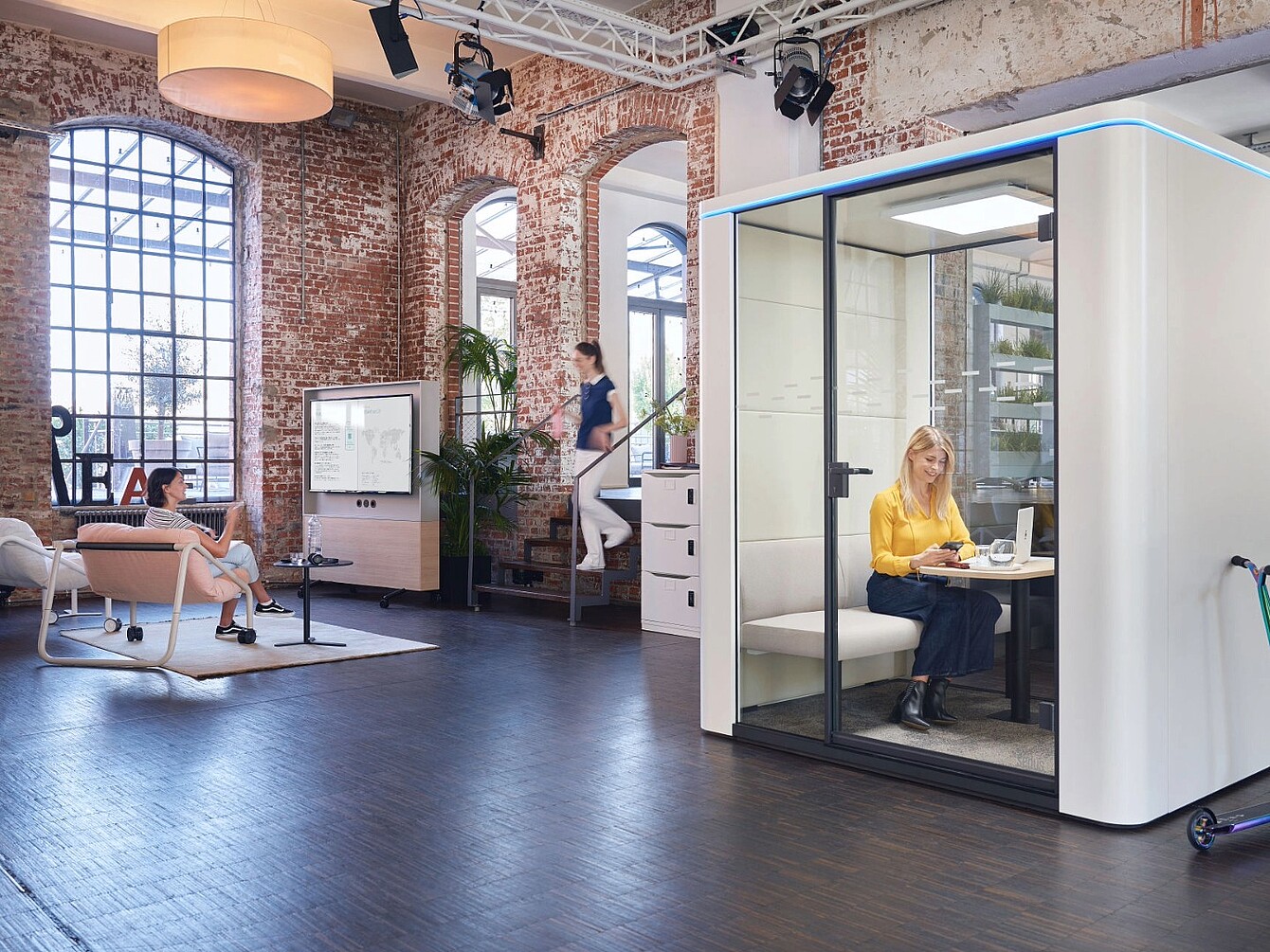 Espacio de oficina moderno con paredes de ladrillo visto y grandes ventanas en el que se ve a una mujer sentada dentro de una cabina acústica acristalada trabajando con un ordenador portátil y a otras dos mujeres en la zona abierta, una sentada en una silla y la otra subiendo las escaleras