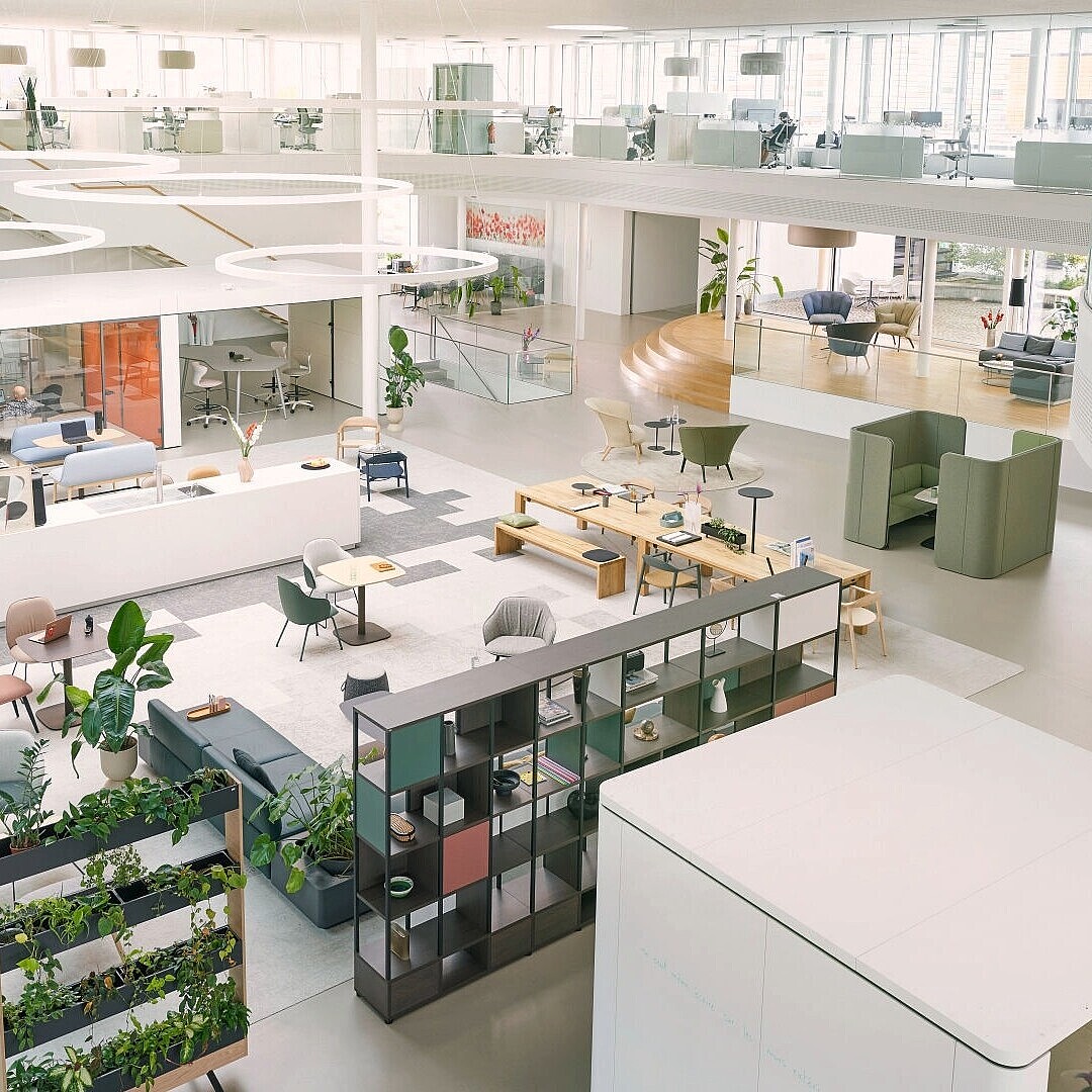 Spacious modern office interior with open seating areas, modular furniture, plants, and large windows providing natural light