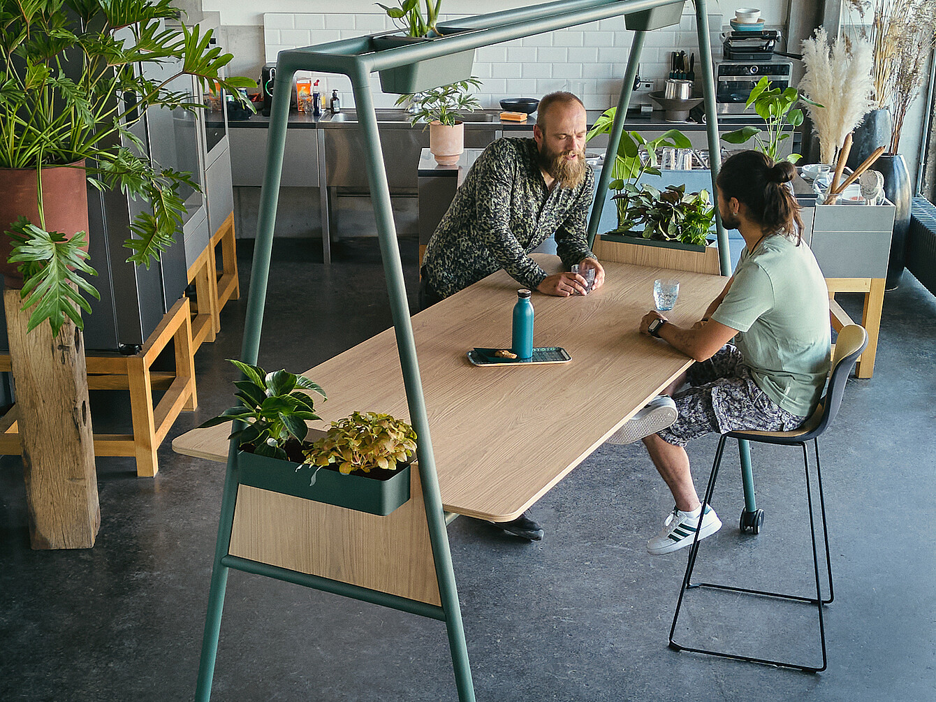 Dos hombres conversando en una mesa de madera con maceteros integrados en un espacio de trabajo moderno diseñado para inspirar el trabajo concentrado y la creatividad colaborativa, rodeados de plantas y elementos de cocina.