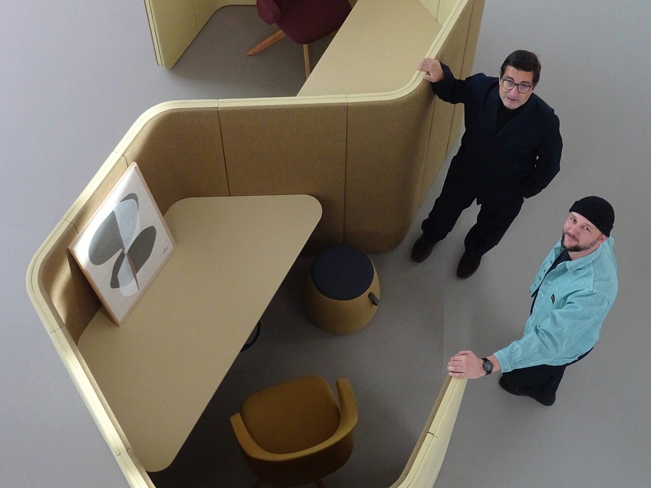 Dos hombres (Rudi Raschke y Konstantin Thomas) están junto a un espacio de trabajo particionado curvo de color beige con una obra abstracta montada, una silla de color mostaza y un pequeño taburete redondo en su interior.