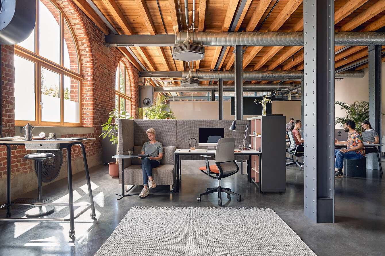 Ufficio open space con travi a vista in legno, grandi finestre ad arco e colonne portanti in metallo, in cui una persona seduta su un divano dallo schienale alto lavora al computer portatile e due persone conversano a un tavolo sullo sfondo
