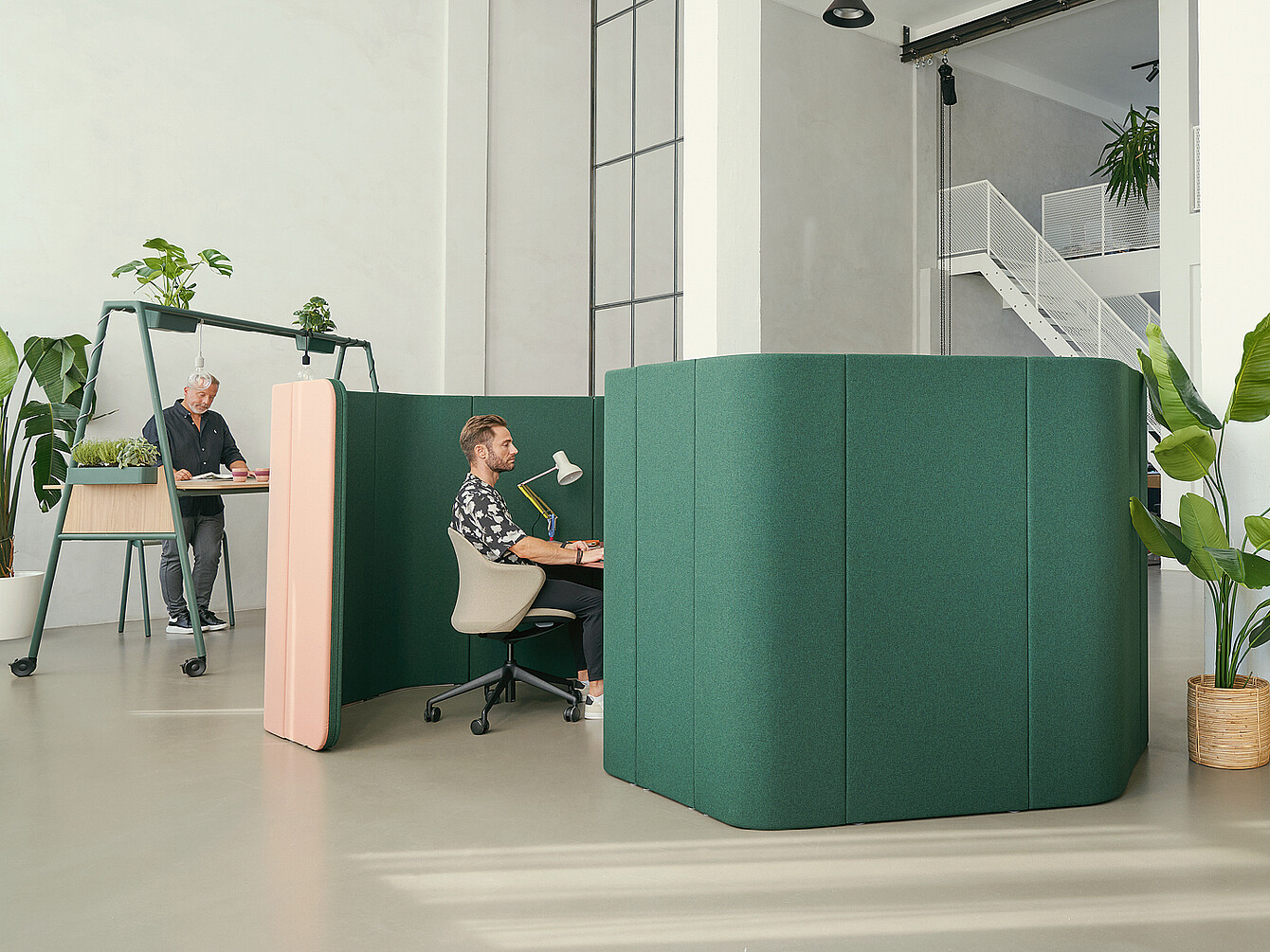 Due postazioni di lavoro in un moderno ufficio con pareti divisorie acustiche verdi. Una persona è seduta a una scrivania con una lampada, mentre un’altra è in piedi davanti a un tavolo più alto con delle piante.