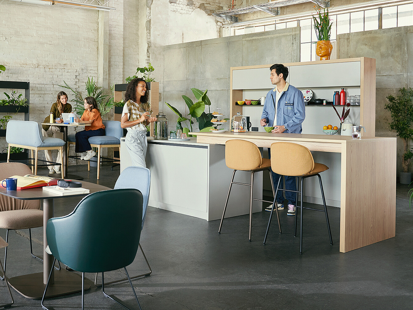 Cafetería de trabajo moderna con asientos flexibles y una barra central donde un hombre con chaqueta azul atiende a una mujer con delantal blanco, rodeados de plantas y paredes de estilo industrial.