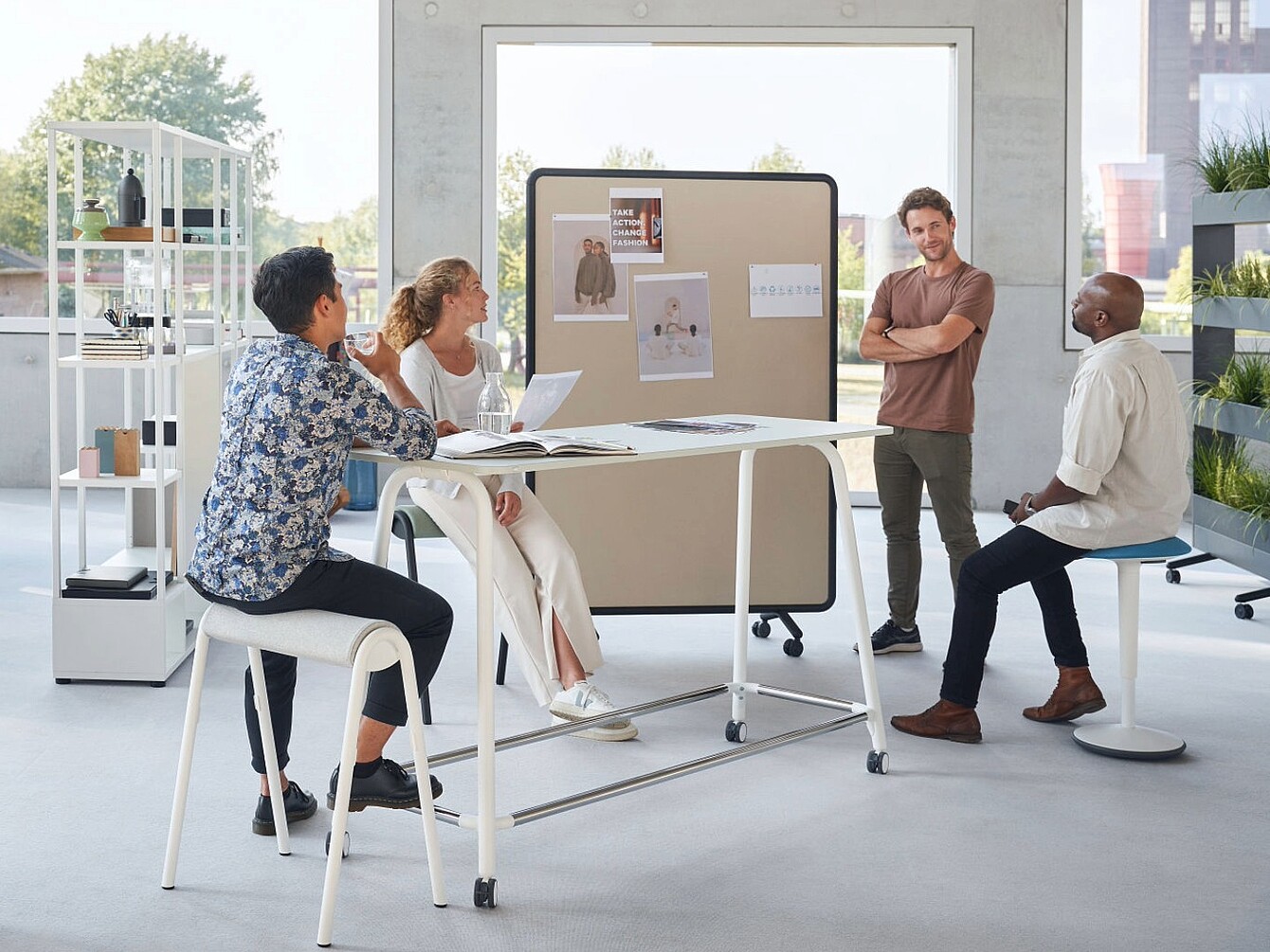 De afbeelding toont een moderne, open kantoorruimte waar meerdere mensen samen aan een hoge tafel werken en documenten bespreken. Verplaatsbare scheidingswanden, rekken en planten helpen de ruimte te organiseren en creëren flexibele zones voor teamwork en samenwerking. Grote ramen laten veel natuurlijk licht binnen, waardoor een lichte en uitnodigende werkomgeving ontstaat.