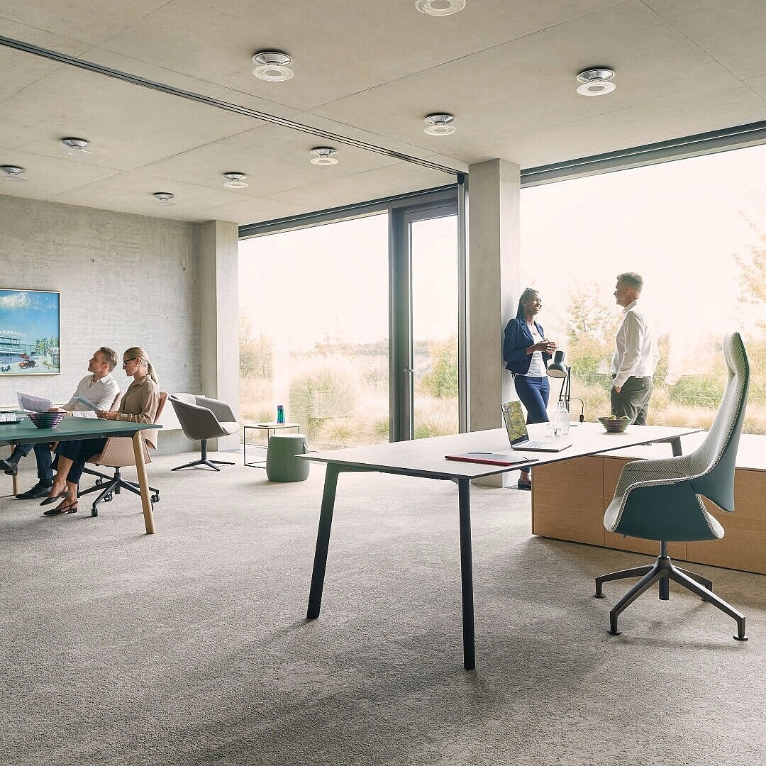 Modernes Büro mit großen Fenstern, mehreren Personen in Besprechung und Stehgespräch, flexiblen Sitzmöglichkeiten und hellem, offenem Raumkonzept