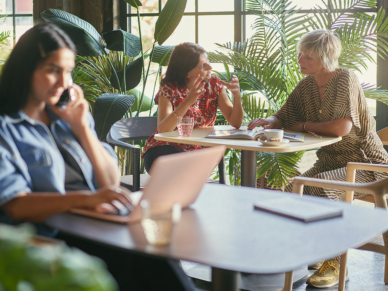 Drei Frauen im Smart Café: Eine arbeitet am Laptop und telefoniert, zwei unterhalten sich lebhaft bei Getränken, umgeben von Pflanzen und natürlichem Licht
