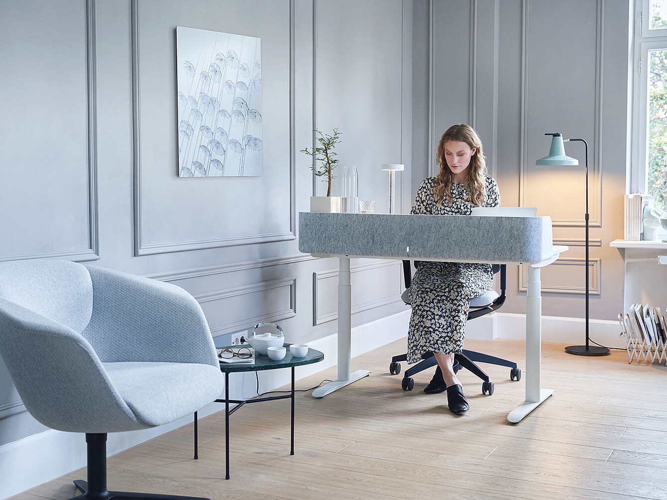Mujer con vestido floral sentada en un moderno escritorio Sedus blanco en una habitación gris claro con un sillón azul y una lámpara de pie.