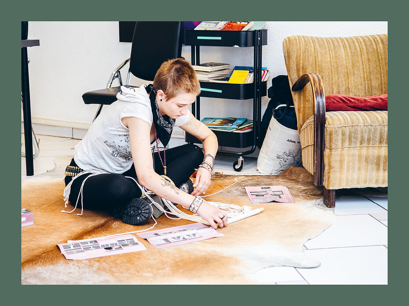 Persona sentada con las piernas cruzadas sobre una alfombra de piel de animal de color marrón claro, ordenando papeles rosas con imágenes en blanco y negro, cerca de un sillón beige y una silla negra en una habitación.