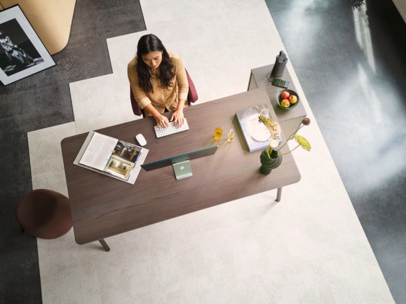 Frau sitzt an einem Schreibtisch und tippt auf einer Tastatur, auf dem Tisch stehen ein Monitor, eine Vase mit Blüten, ein Buch, ein Glas Orangensaft und eine Obstschale