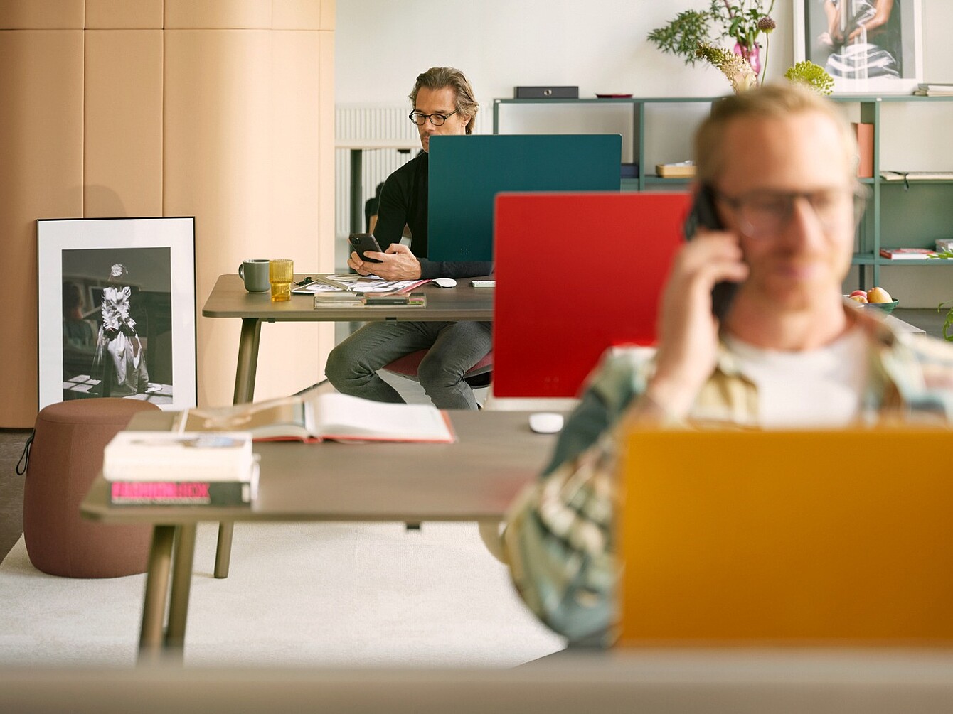 Due collaboratori lavorano alle scrivanie con pannelli divisori colorati, mentre una persona in primo piano parla al telefono.