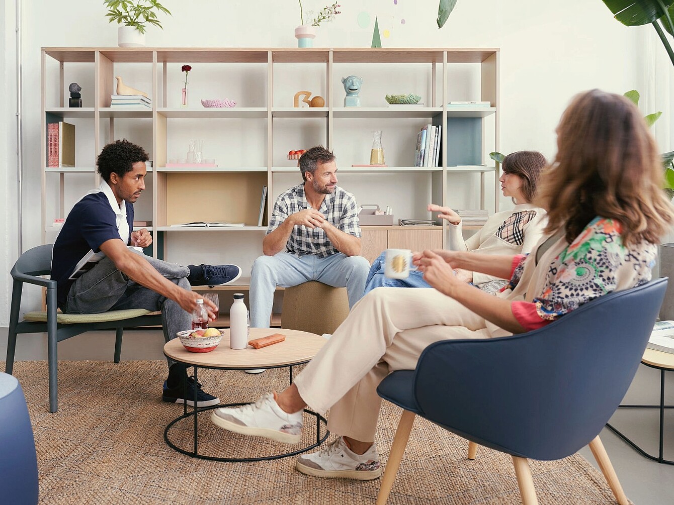 Quattro persone sedute in un’area lounge con sedie moderne e un tavolino rotondo davanti a una grande scaffalatura con oggetti decorativi e libri
