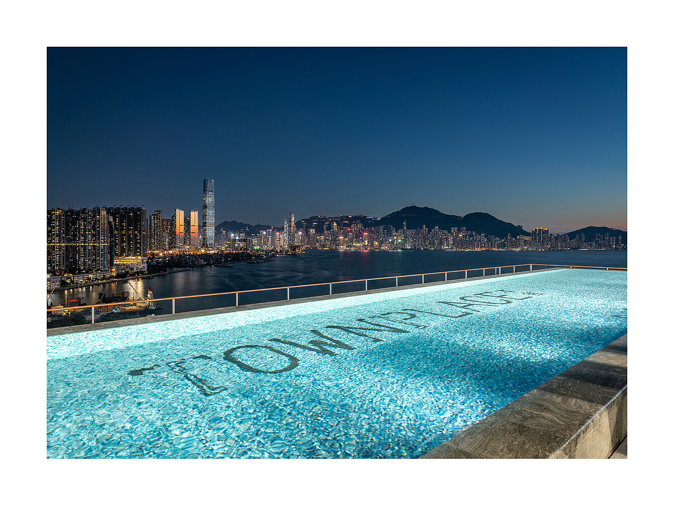 Piscine à débordement sur le toit avec le mot « TOMMY LIFE » visible au fond de la piscine, surplombant la silhouette de la ville au crépuscule 