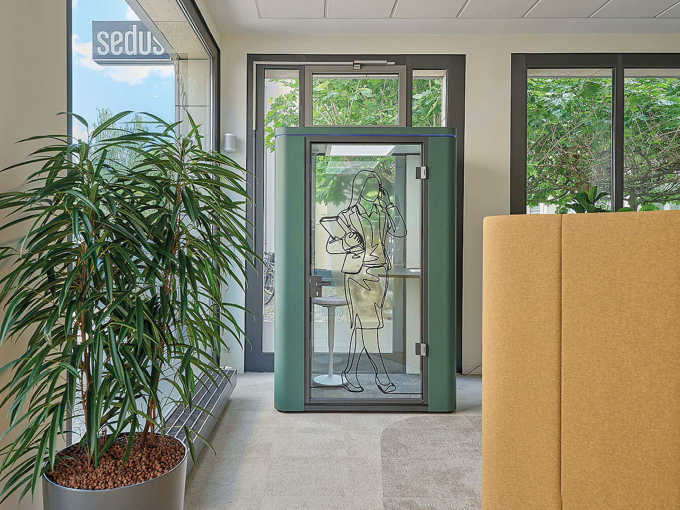 Piccola cabina insonorizzata verde con porta in vetro con disegno di una donna che tiene un telefono, collocata in un moderno spazio ufficio vicino a grandi finestre e a una pianta alta in vaso.