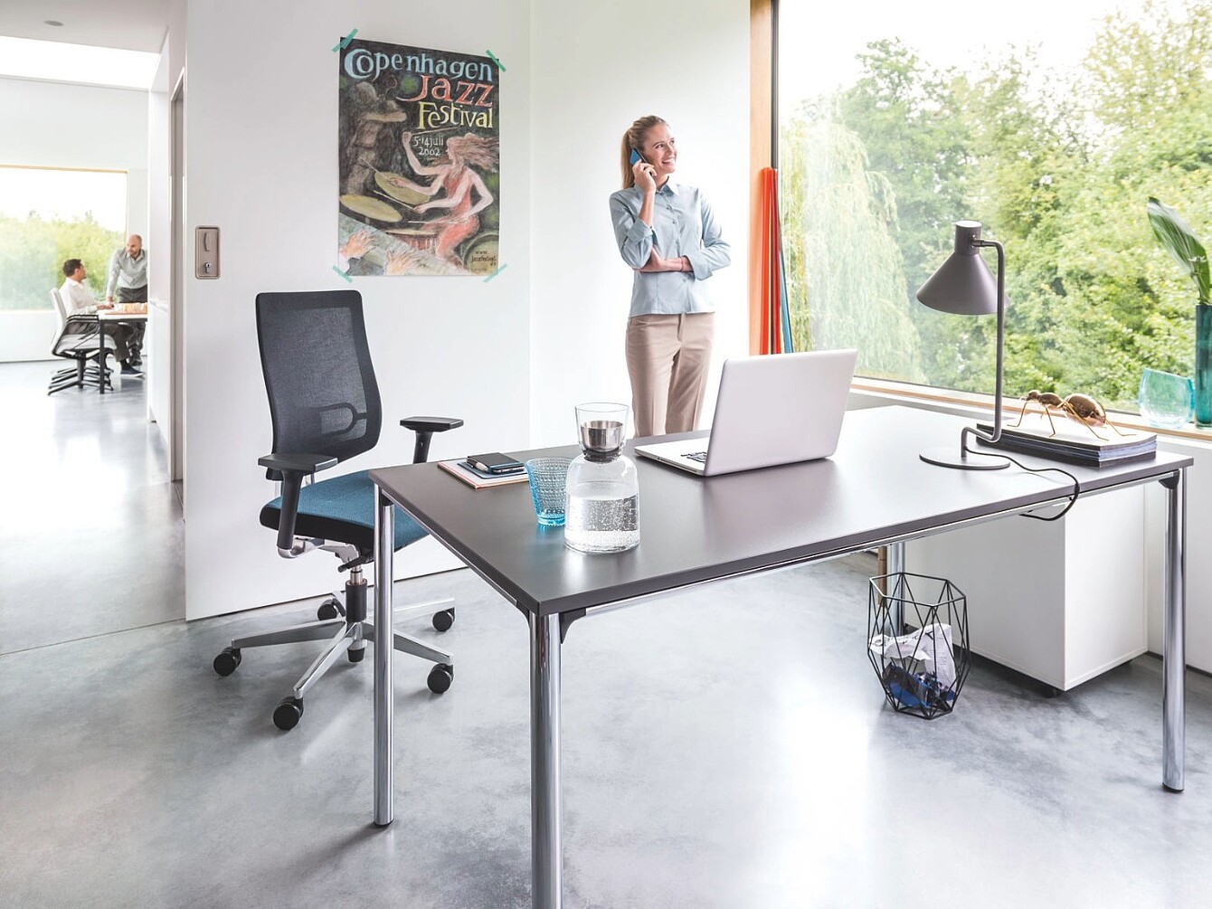 Moderner Büroarbeitsplatz mit Lordosenstütze im ergonomischen Bürostuhl, Schreibtisch mit Laptop, Wasserflasche und Schreibtischlampe, im Hintergrund steht eine Frau am Fenster und telefoniert