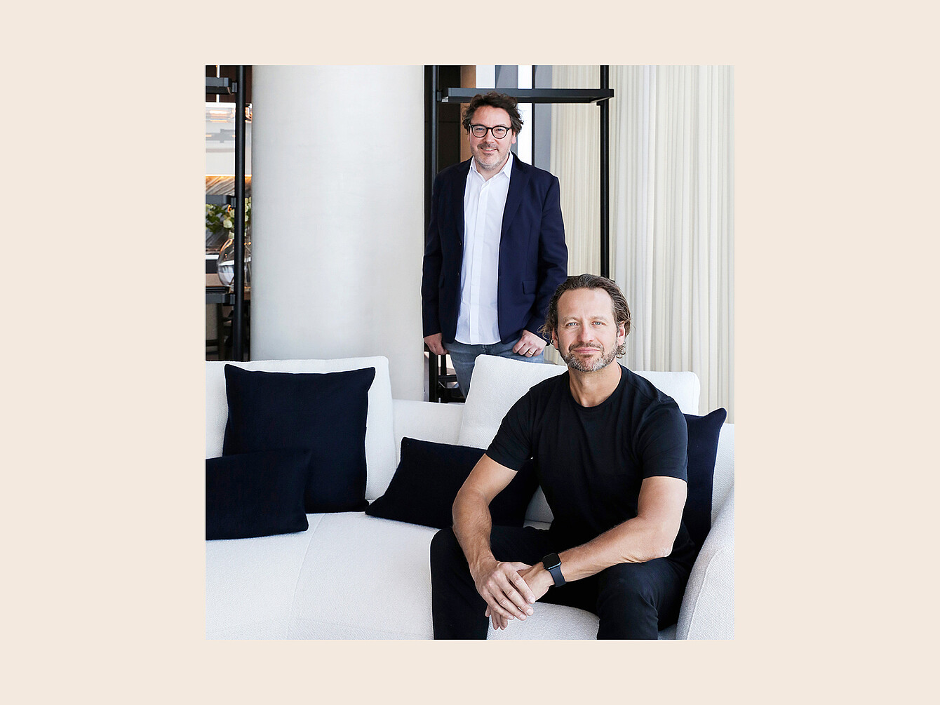 Deux hommes dans un intérieur moderne avec un canapé blanc et des coussins noirs, l’un assis et l’autre debout derrière le canapé.