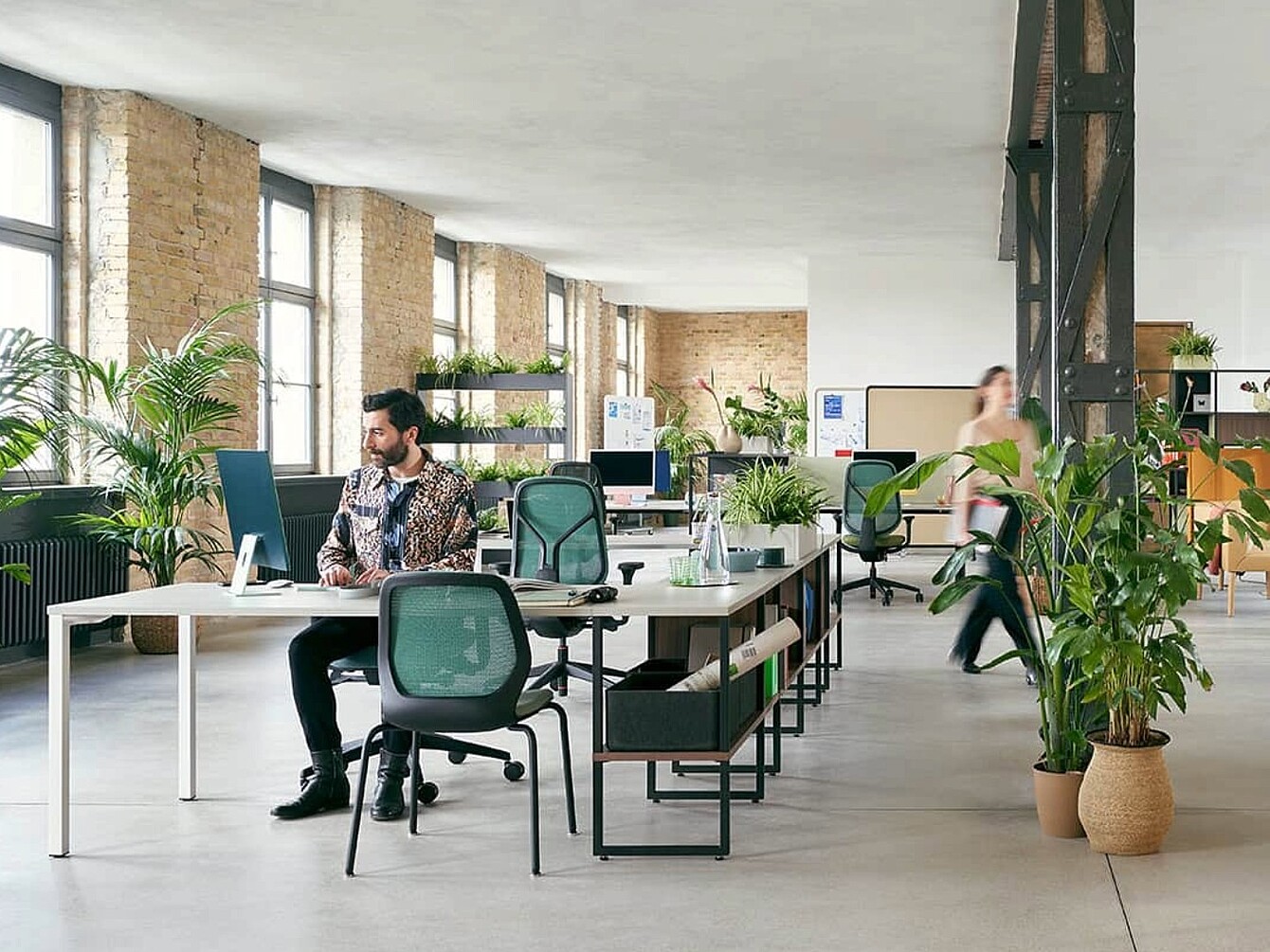 Spazio ufficio aperto e luminoso con diverse scrivanie, sedie ergonomiche da ufficio, scaffali e numerose piante.