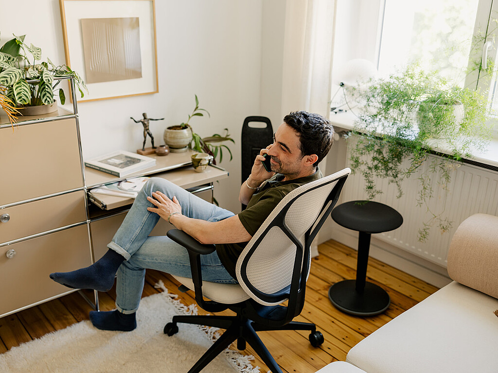 Person sitting on an ergonomic office chair with mesh backrest, talking on the phone in a bright home office with plants and wooden flooring.