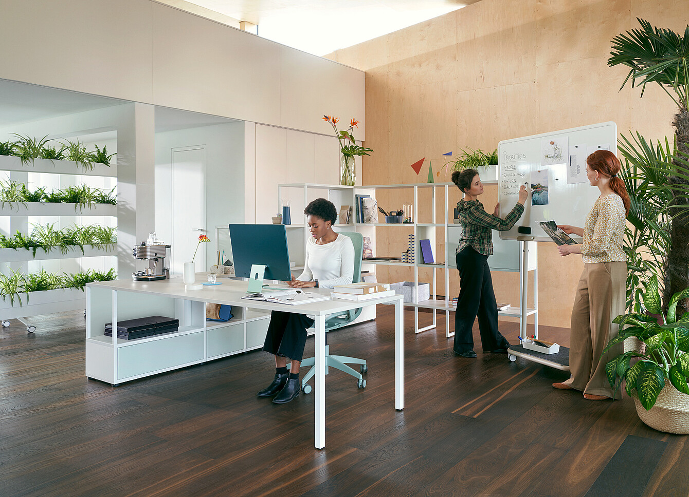 Tre donne in un ufficio moderno con piante: una seduta alla scrivania che lavora al computer e due in piedi accanto a una lavagna che discutono di appunti