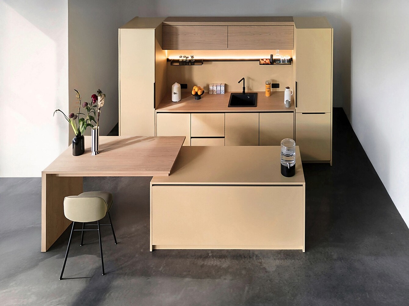 Allestimento minimalista del se:café bar con armadi alti beige, lavello integrato, illuminazione sotto i mobili, prolunga del piano di lavoro in legno con sgabello e vasi decorativi con fiori