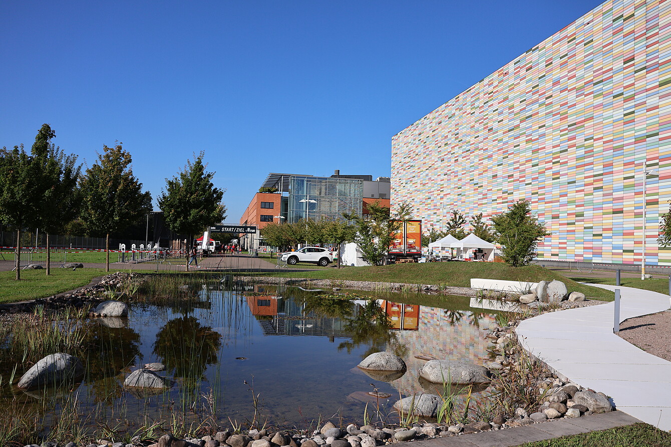 Teich mit Felsen und Pflanzen vor einem farbenfrohen, gekachelten Gebäude und einem modernen Glasgebäude, mit weißen Zelten und geparkten Autos in der Nähe unter einem strahlend blauen Himmel.