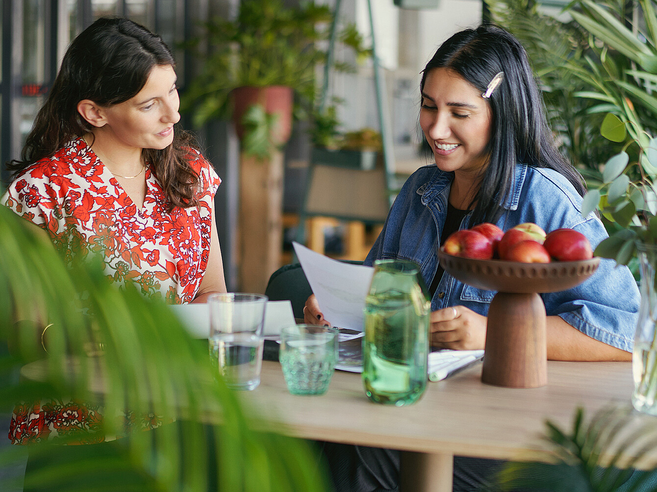 Dos mujeres conversan amistosamente sobre unos documentos en una mesa de madera, en un luminoso espacio de oficina con plantas y un cuenco de manzanas, que ilustra las visitas intencionadas a la oficina para una interacción y un apoyo significativos.