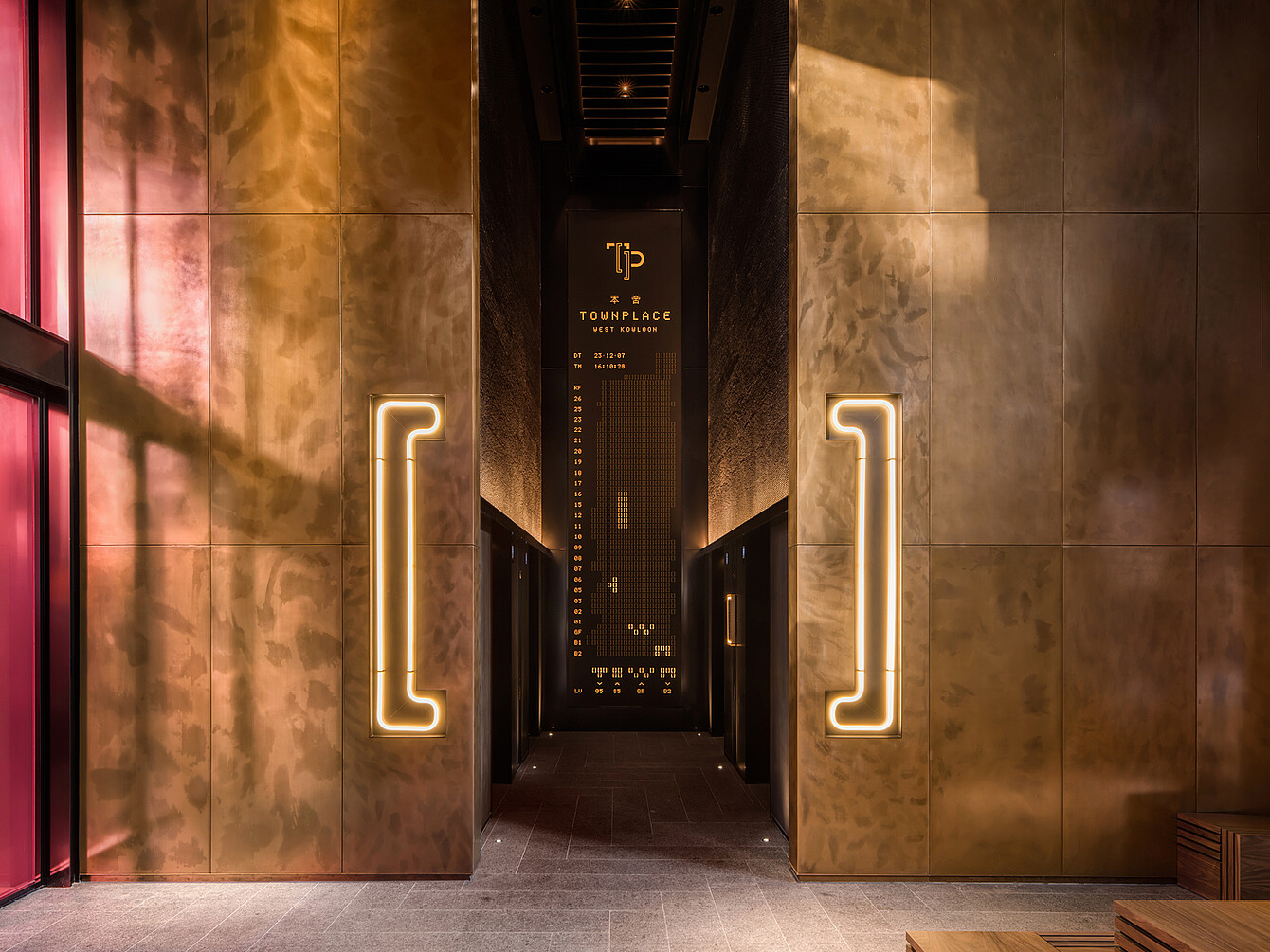 Couloir étroit avec des murs couleur bronze et des poignées verticales éclairées de chaque côté menant à une entrée sombre avec une enseigne indiquant « TP TOWNPLACE ». 