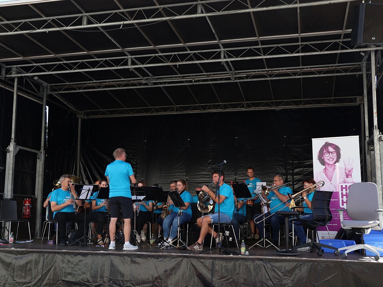 Group of musicians in blue shirts seated on a stage playing brass and woodwind instruments with a conductor standing in front