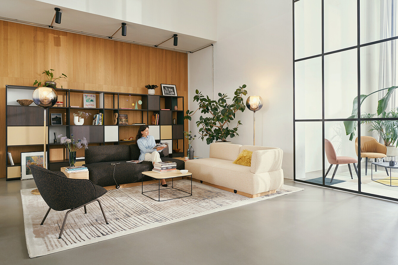Woman reading in a quiet lounge area with sofas, shelving, plants and warm wooden surfaces.