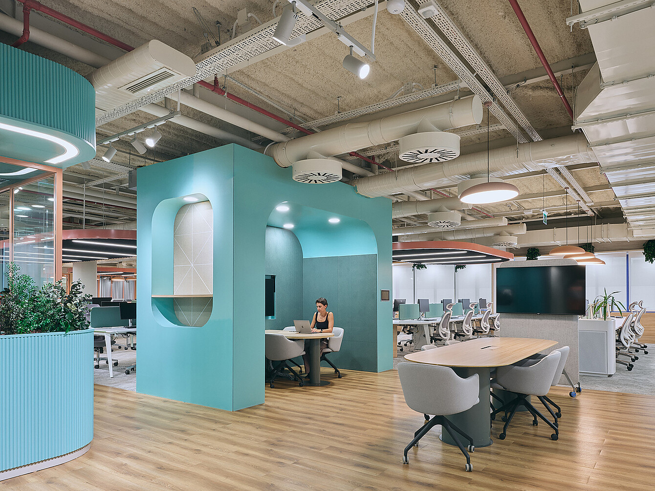 Espacio de oficina diáfano con suelo de madera clara, una cabina de reuniones cerrada de color turquesa con una persona trabajando en un portátil en su interior, varias sillas de oficina blancas, escritorios con monitores de ordenador y tuberías y conductos a la vista en el techo