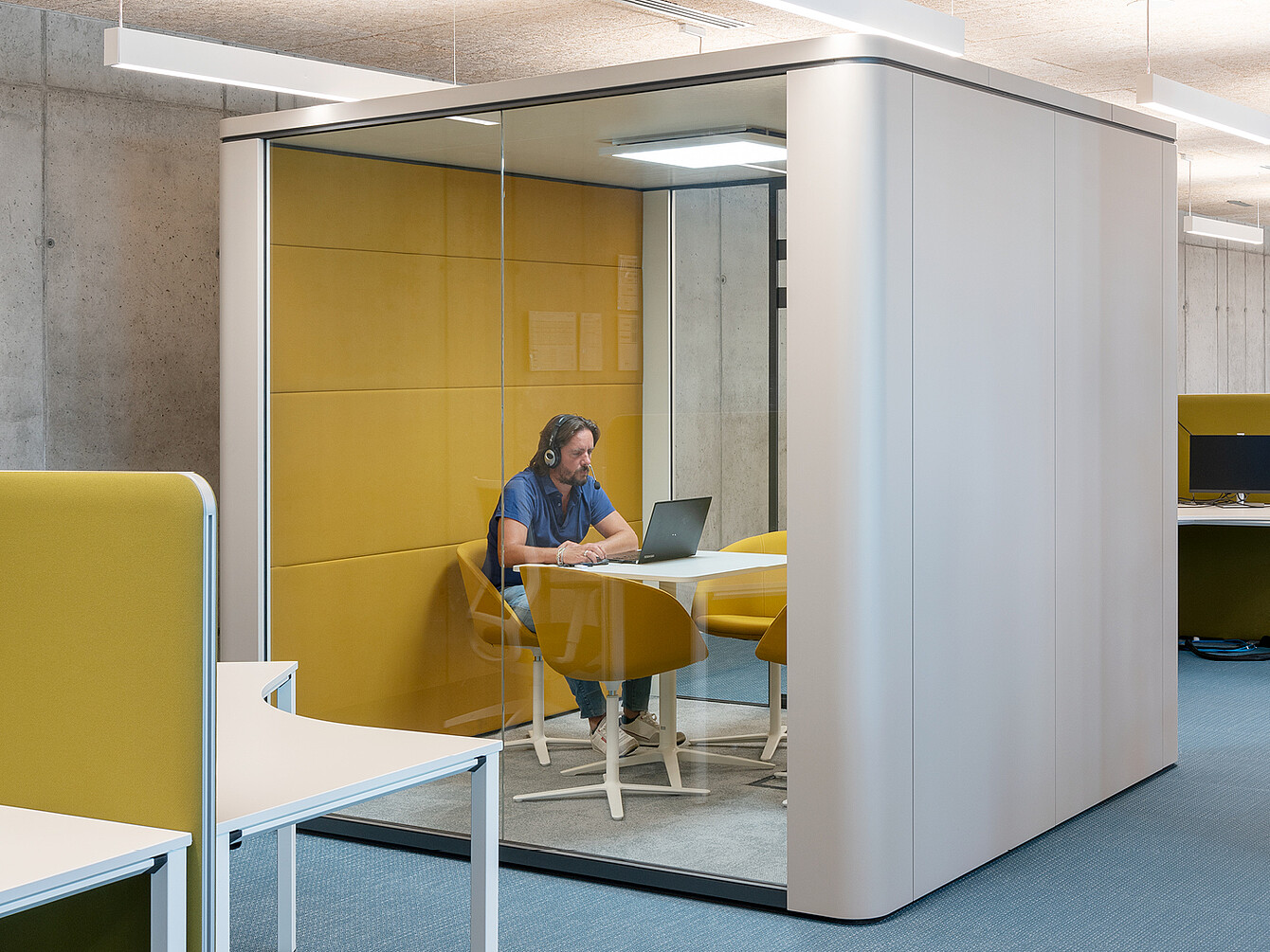 Persona con auriculares sentada en una mesa con un ordenador portátil dentro de una cabina de oficina con paredes de cristal y paredes y sillas acolchadas de color amarillo