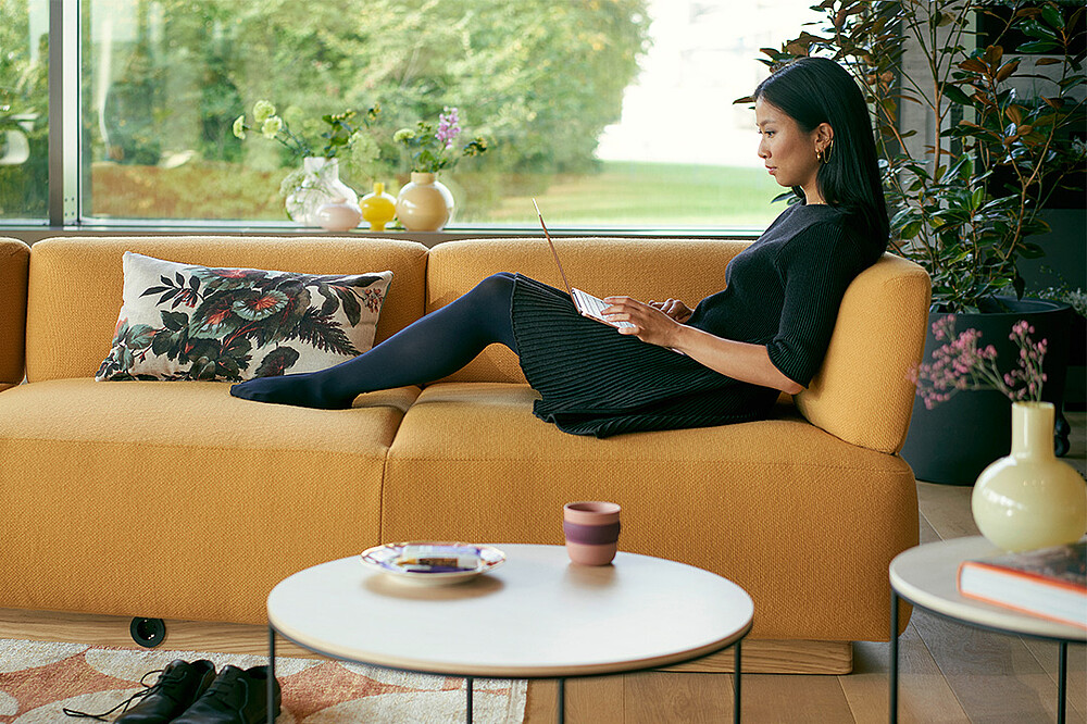 Eine Frau sitzt entspannt auf einem se:living von Sedus und arbeitet mit einem Laptop, der auf ihren Oberschenkeln ruht. Im Vordergrund ist unscharf ein se:living side table zu erkennen, der die gemütliche und flexible Atmosphäre des Raumes unterstreicht. Im Hintergrund dominiert ein großes Fenster, das den Raum mit natürlichem Licht erfüllt und einen weiten Blick auf den Garten bietet. Diese Szene veranschaulicht das Konzept des duktilem Raumes, der als flexibler Knotenpunkt in hybriden Büros dient und individuelles Arbeiten sowie den Übergang zu dezentralen Arbeitsmethoden unterstützt.