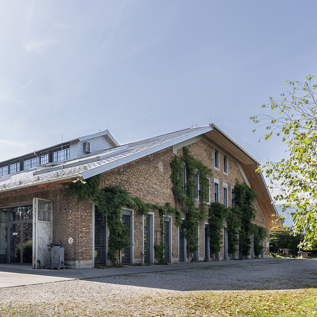 Langes, zweigeschossiges Backsteingebäude mit großen Fenstern und begrünten Fassaden in einem von Bäumen und Sträuchern umgebenen Co-Working-Space