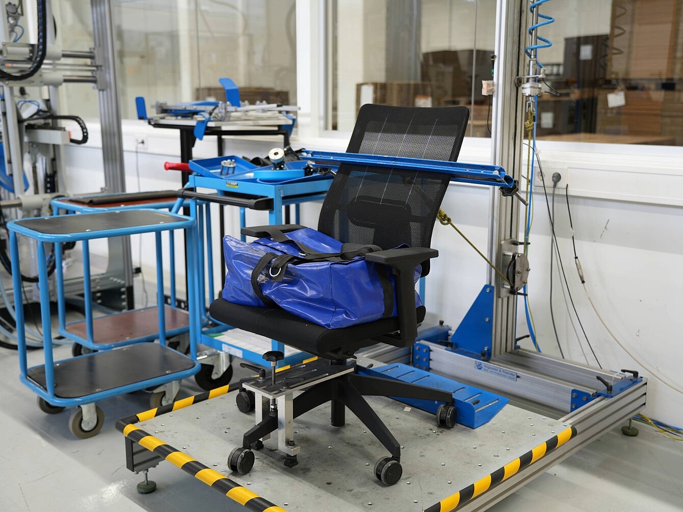 Silla de oficina fijada en un bastidor de ensayo metálico azul dentro de un laboratorio, con equipos y carros al fondo