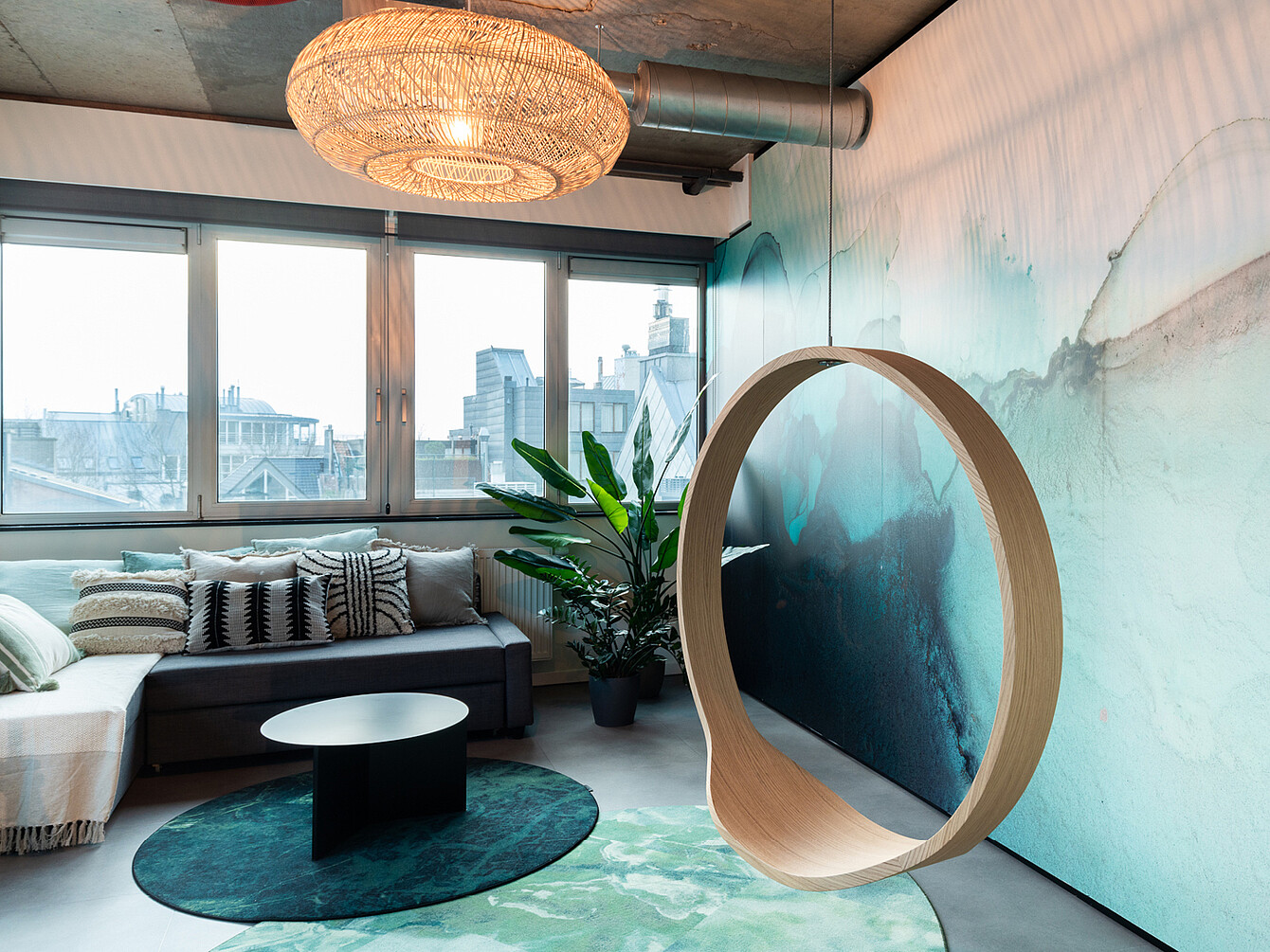 Moderner Lounge-Bereich mit grauem Sofa, rundem schwarzen Tisch, grünem Teppich, großer Pflanzen und hängendem runden Holzobjekt vor einer abstrakten Wandgestaltung mit Stadtfenstern im Hintergrund