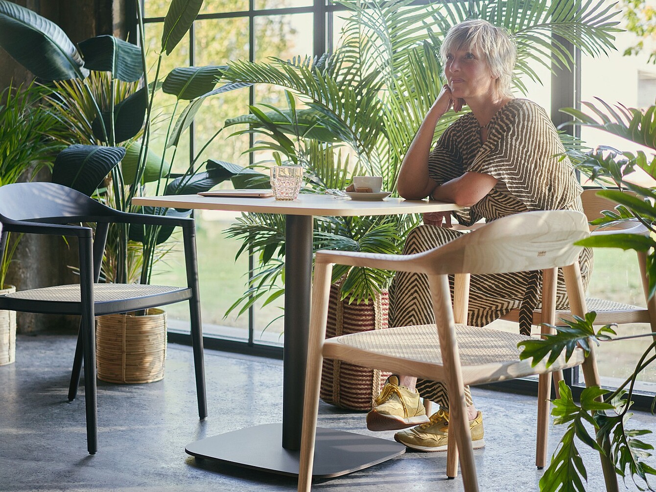 Personne assise à une table dans une pièce dotée de grandes fenêtres et entourée de nombreuses plantes de bureau