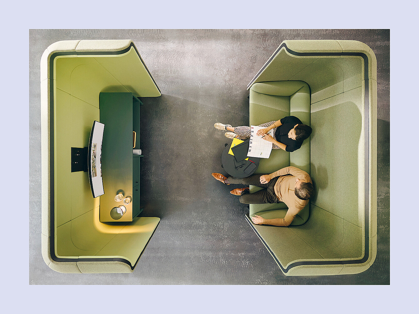 Dos personas sentadas en una cabina verde de respaldo alto con una pequeña mesa redonda en la que hay papeles y una bolsa entre ellas, frente a una cabina a juego con una pantalla curva montada y estantes