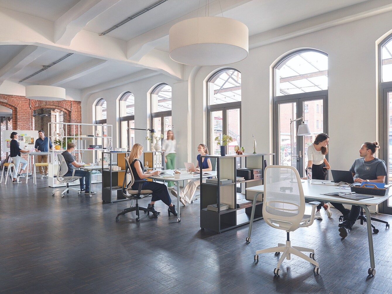 Ufficio open space con persone al lavoro alle scrivanie e grandi finestre ad arco lungo una parete