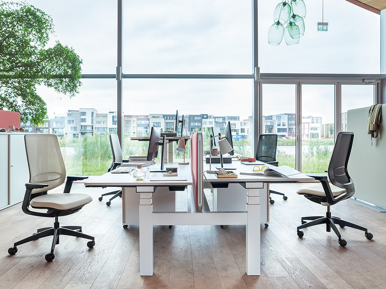 Moderno espacio de trabajo con cuatro escritorios dispuestos en dos filas enfrentadas, sillas ergonómicas, monitores de ordenador y grandes ventanas con vistas al exterior de la ciudad...