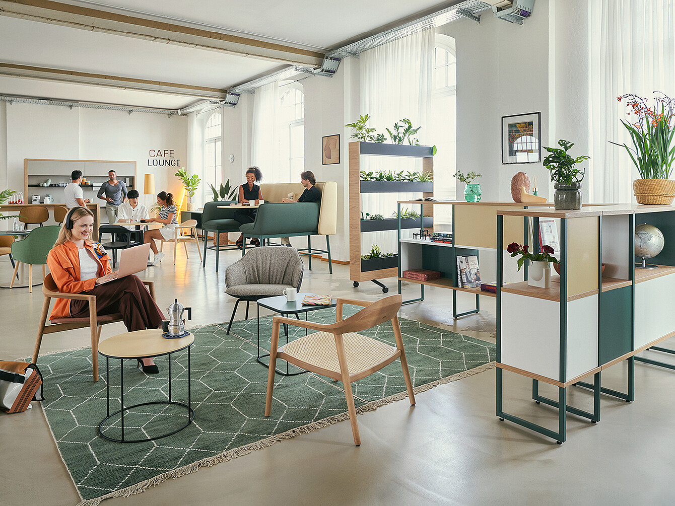 Un moderno Work Café con varie aree salotto con piante. In primo piano, una donna con una giacca arancione è seduta su una sedia con un laptop. Sullo sfondo, alcune persone conversano e lavorano ai tavoli. Lo spazio include scaffali con oggetti decorativi e un cartello con la scritta “CAFE LOUNGE”.