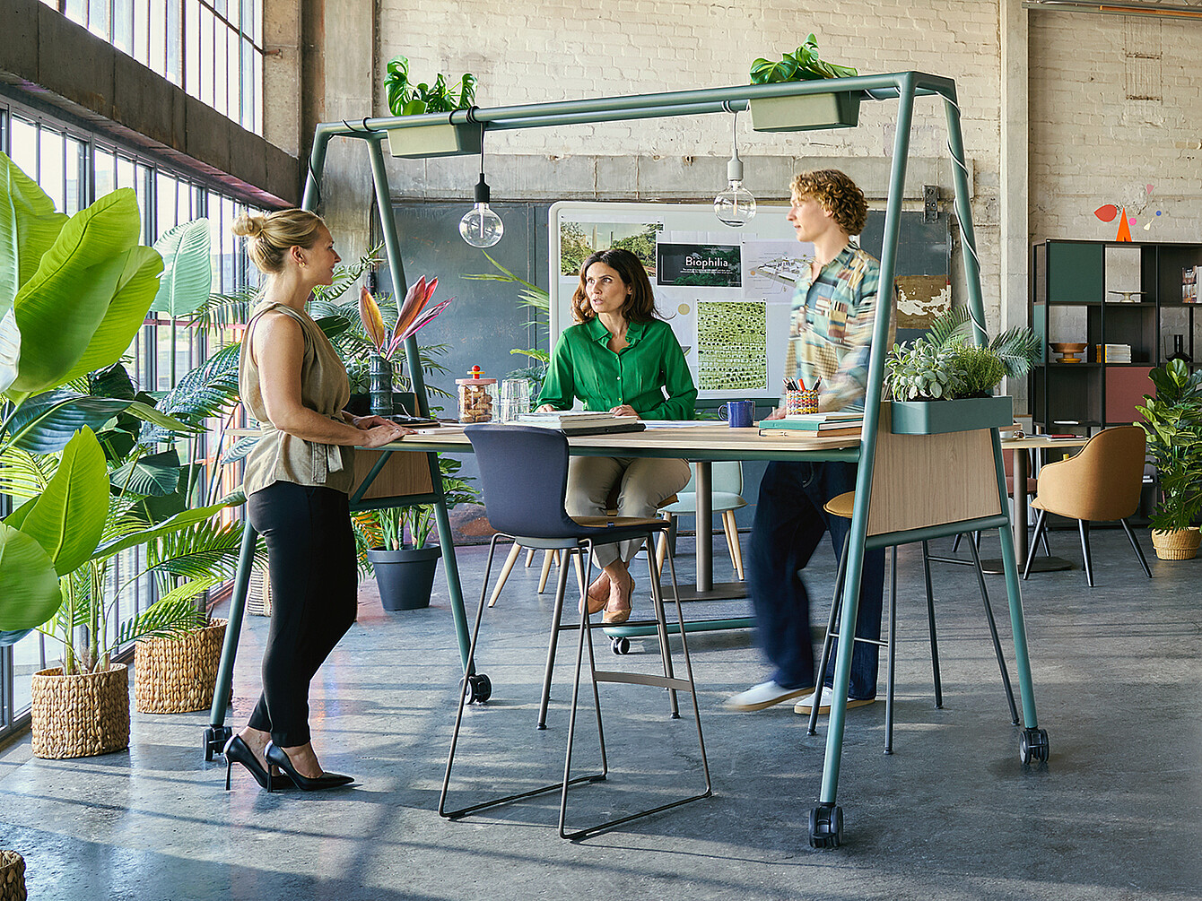 Tres compañeros colaboran alrededor de una mesa alta con plantas en una oficina luminosa y llena de vegetación, ejemplificando el diseño biofílico del Garden Work Café.