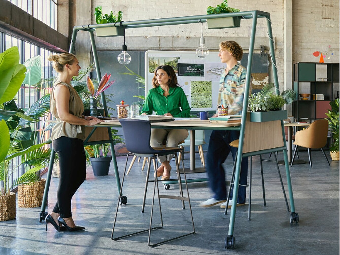 Trois personnes autour d’une table haute surmontée et entourée de plantes, dans un bureau spacieux doté de grandes fenêtres et de murs en briques apparentes